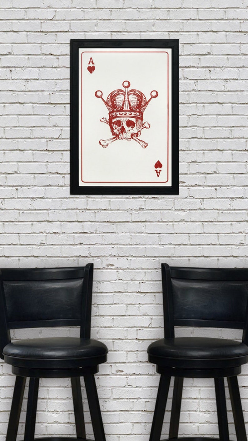 May include: A framed art print featuring a red skull wearing a crown, crossed bones, and the Ace of Spades and Hearts symbols. The artwork is displayed on a white brick wall, with two black chairs below.