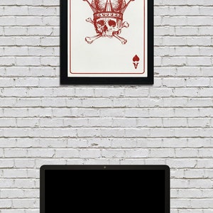 May include: A framed art print featuring a red and white Ace of Hearts card design. The card depicts a skull wearing a crown, with crossbones, and the Ace symbol. The print is in a black frame, hanging on a white brick wall.