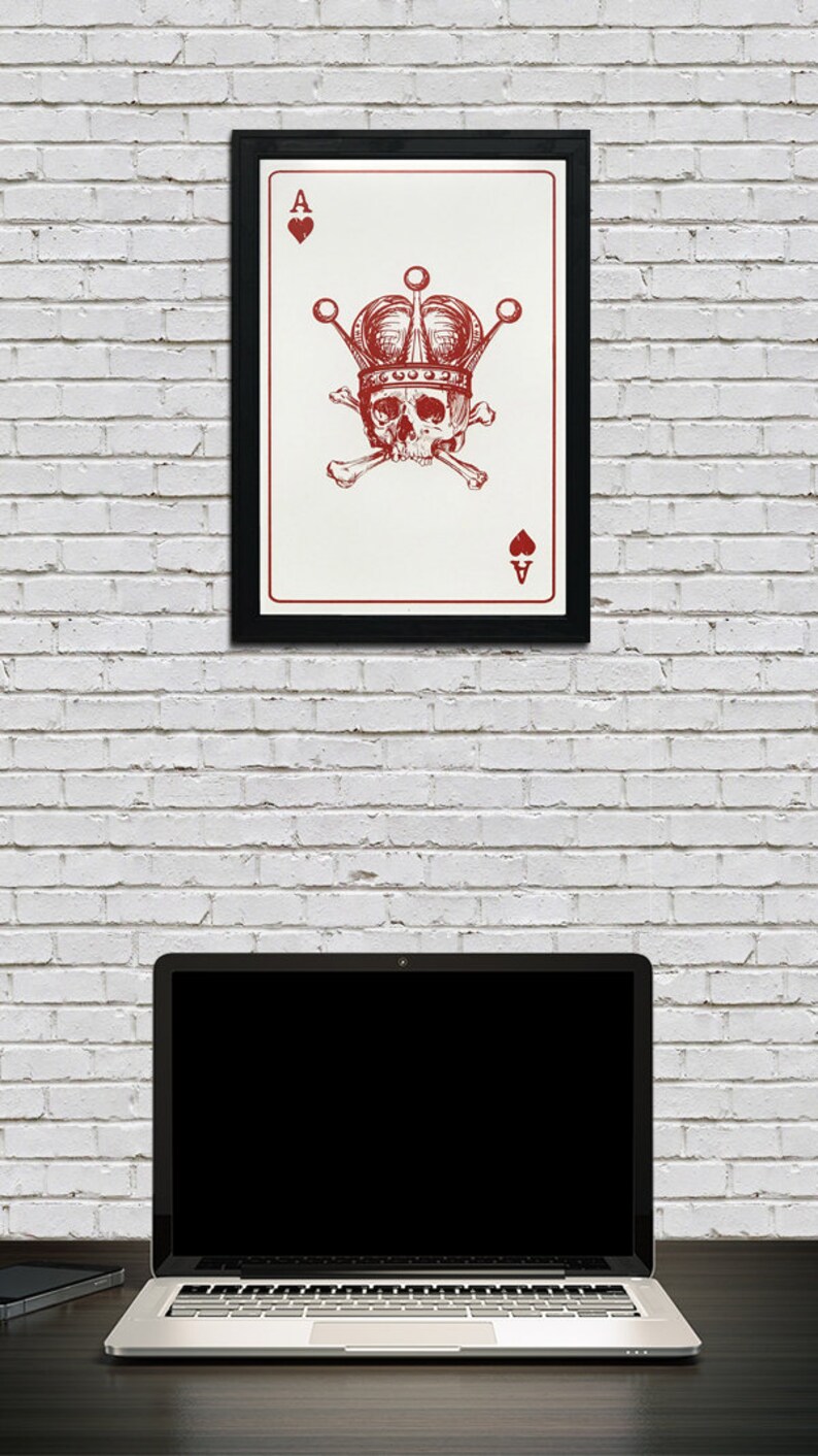 May include: A framed art print featuring a red and white Ace of Hearts card design. The card depicts a skull wearing a crown, with crossbones, and the Ace symbol. The print is in a black frame, hanging on a white brick wall.