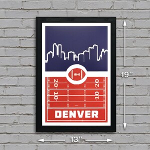 May include: A black framed print with a red, white, and blue design. The print features a skyline of Denver, Colorado, with a football field in the foreground. The text "DENVER" is printed in white on the red field. The print measures 13 inches wide by 19 inches tall.