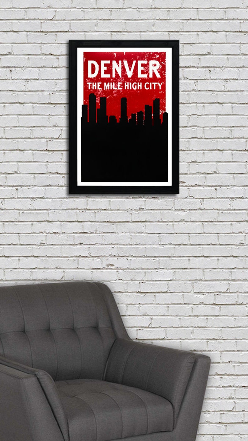 May include: A framed art print featuring the Denver skyline in black against a red background. The word "DENVER" is displayed in white, above "THE MILE HIGH CITY". The print is in a black frame, hanging on a white brick wall.
