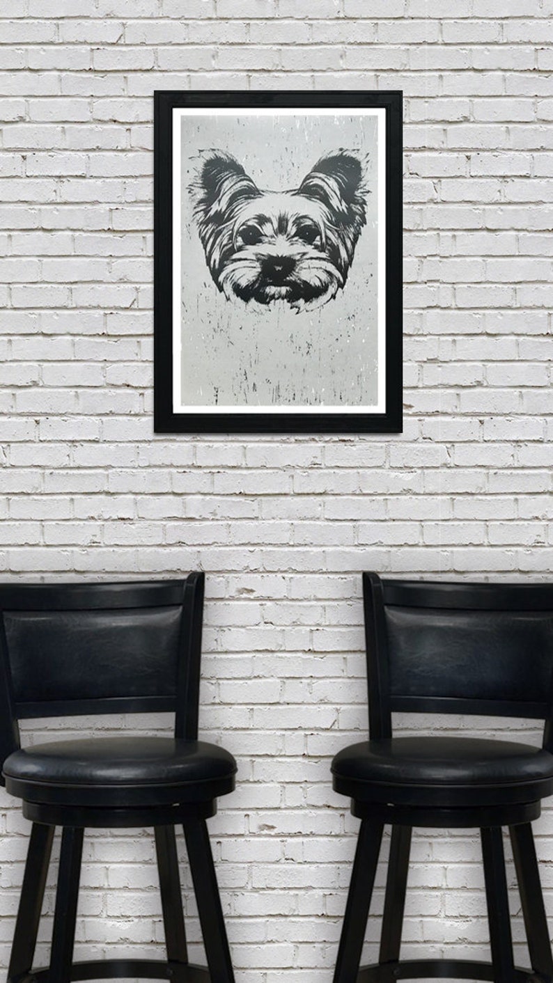 May include: A black and white framed print of a dog's face. The dog has long fur and is looking directly at the camera. The print is hanging on a white brick wall between two black bar stools.