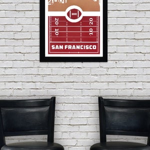 May include: A framed print of a white silhouette of the San Francisco skyline against a brown background. The skyline is set against a red football field with yard lines and a football in the center. The text "SAN FRANCISCO" is printed below the field.
