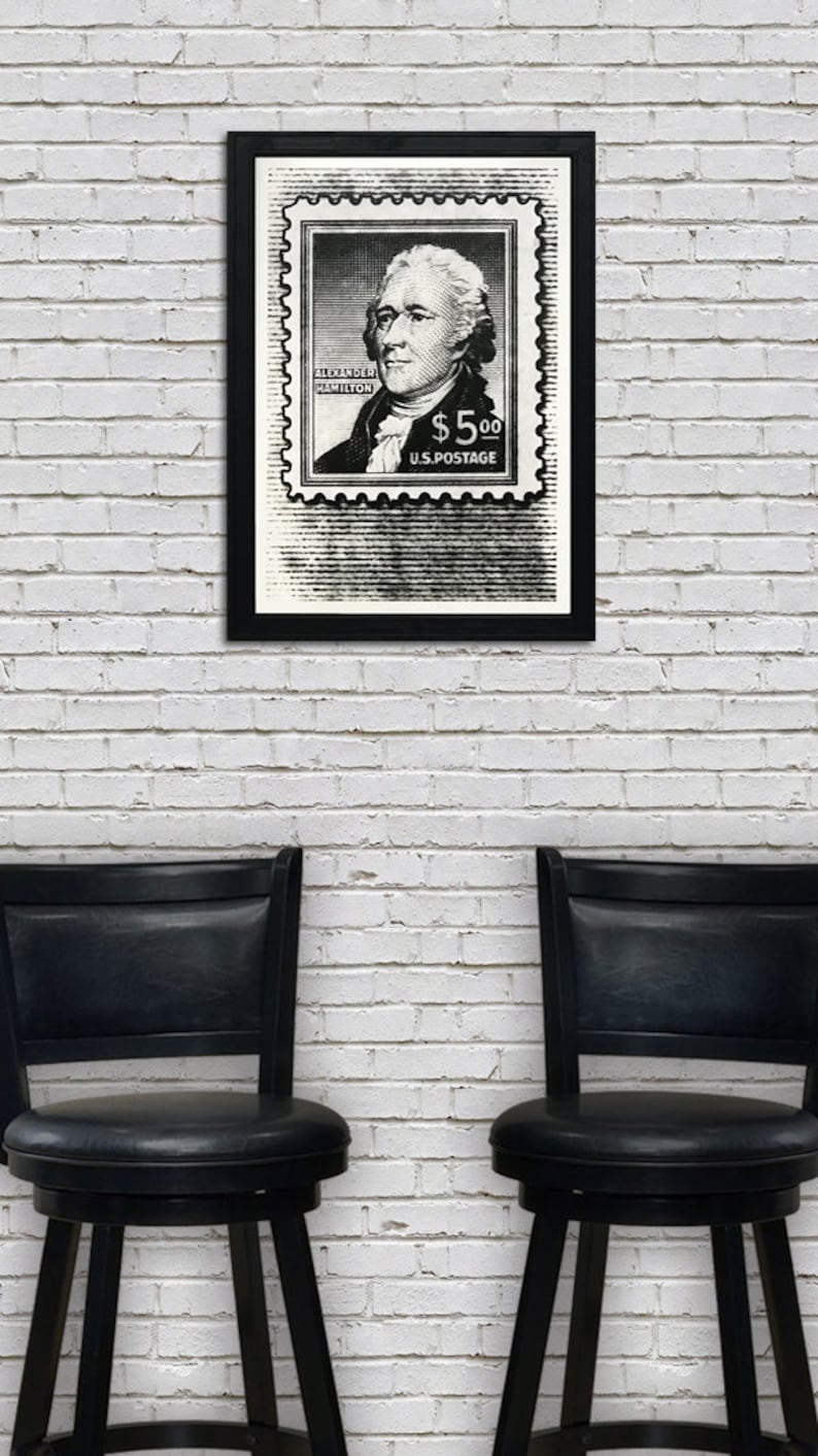 May include: A black and white framed print of a vintage postage stamp featuring a portrait of Alexander Hamilton with the text "ALEXANDER HAMILTON $5.00 U.S. POSTAGE". The print is hanging on a white brick wall between two black bar stools.