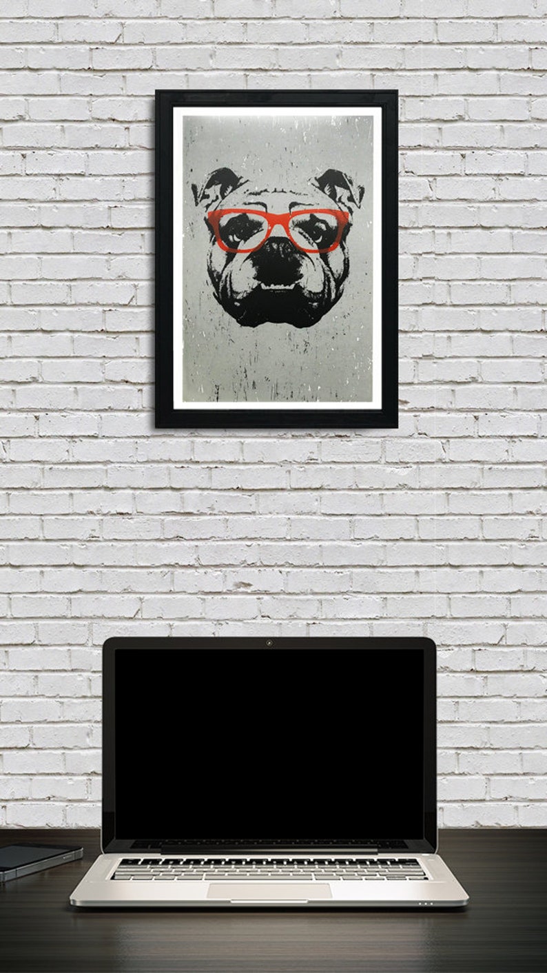 May include: A black and white illustration of a bulldog wearing red glasses. The illustration is framed in a black frame and hanging on a white brick wall. A laptop computer is sitting on a dark surface in front of the wall.