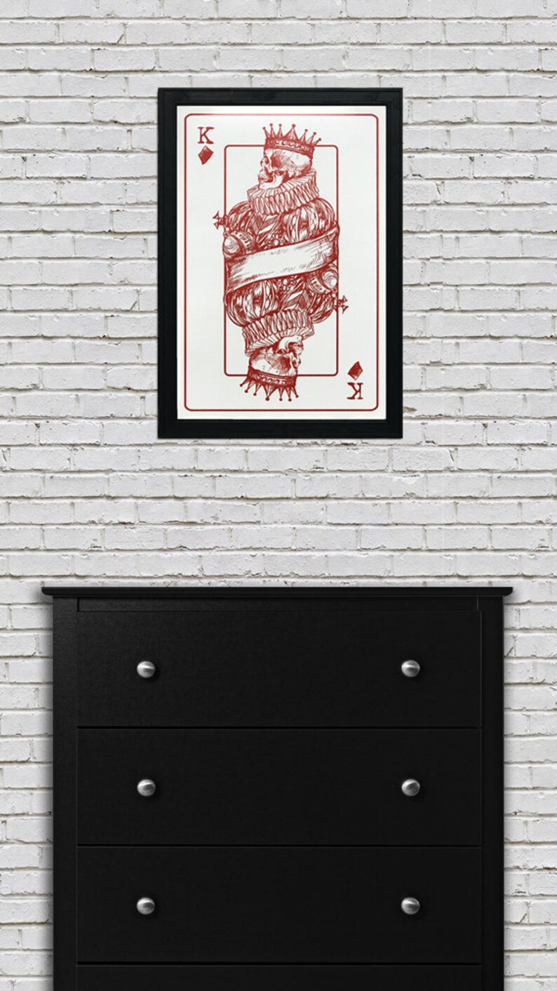 May include: A framed print of a playing card featuring a red skull king of diamonds. The skull is wearing a crown and is surrounded by a banner.