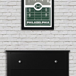 May include: A framed print of a green football field with a white outline of the Philadelphia skyline above it. The text "PHILADELPHIA" is printed below the field.
