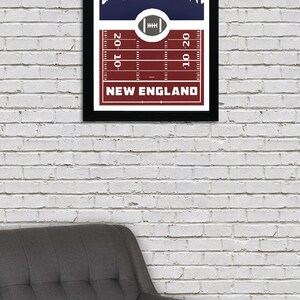 May include: A framed print with a navy blue background featuring a white silhouette of a city skyline. The bottom of the print has a red and white football field design with the text "NEW ENGLAND" and yard lines marked "20", "10", and "10".
