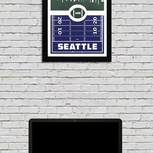 May include: A framed print of a football pitch with a skyline of Seattle, Washington. The pitch is blue and the skyline is green. The text "Seattle" is printed below the pitch. The text "20-10" is printed on the left side of the pitch and the text "10-20" is printed on the right side of the pitch.