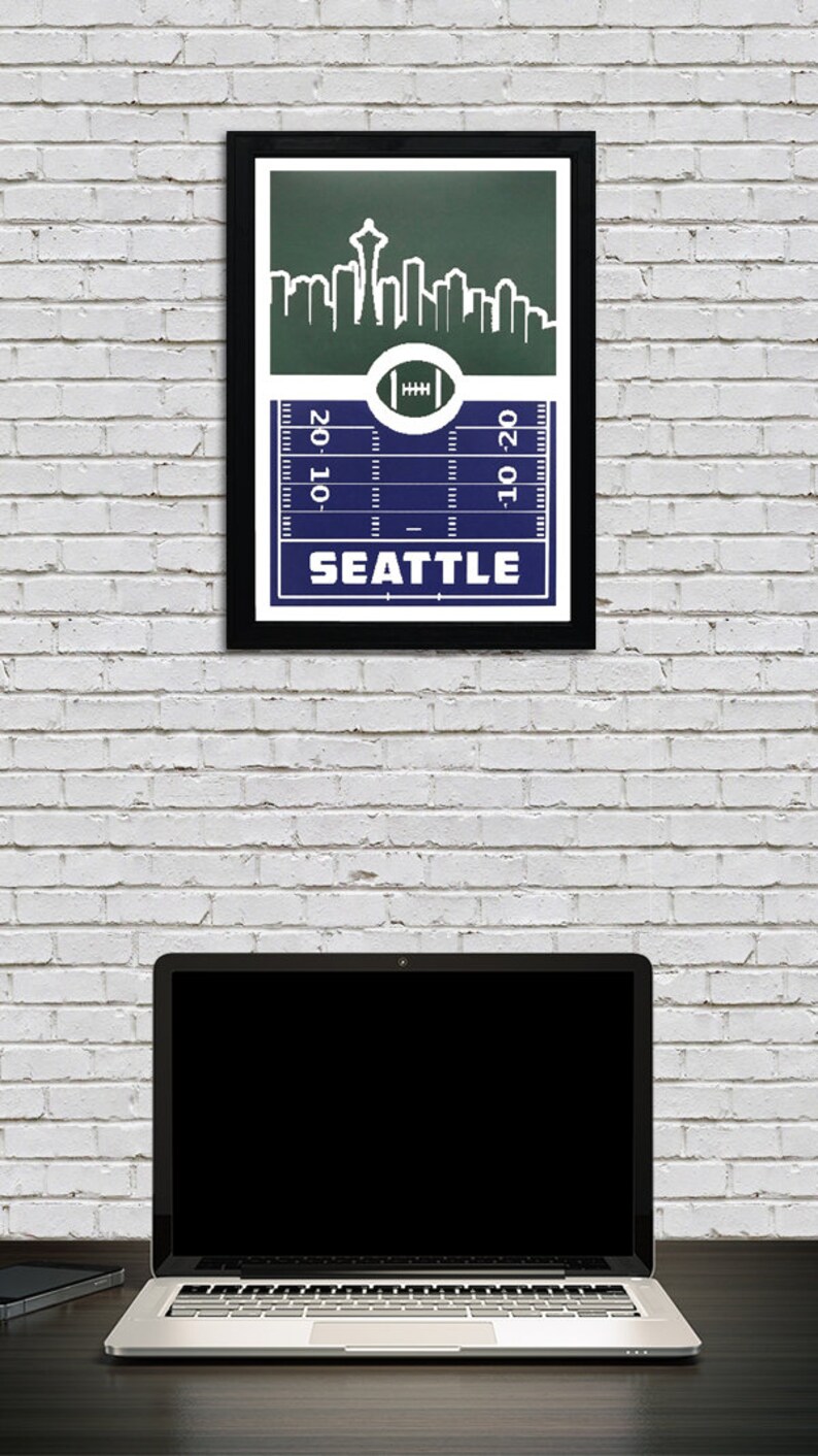May include: A framed print of a football pitch with a skyline of Seattle, Washington. The pitch is blue and the skyline is green. The text "Seattle" is printed below the pitch. The text "20-10" is printed on the left side of the pitch and the text "10-20" is printed on the right side of the pitch.