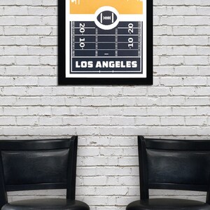 May include: A black framed print with a gold background featuring a white silhouette of the Los Angeles skyline. The bottom of the print shows a black and white football field with the text "LOS ANGELES" and the numbers "20-10" and "10-20".