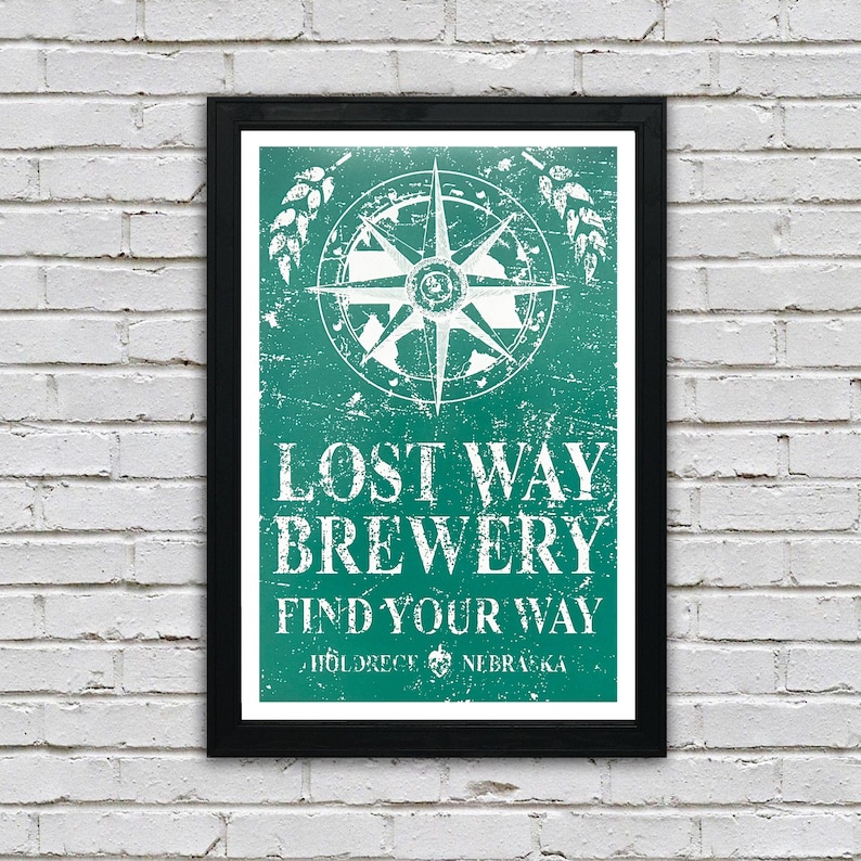 May include: A framed poster with a distressed green background featuring a white compass with a star in the center. The text "Lost Way Brewery Find Your Way" is written in white with a distressed font. The text "Hildreth, Nebraska" is written in white at the bottom of the poster.