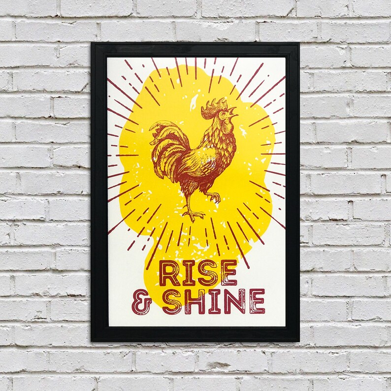 May include: A framed print with a yellow sunburst design and a red rooster illustration. The text "Rise & Shine" is printed in red below the rooster.