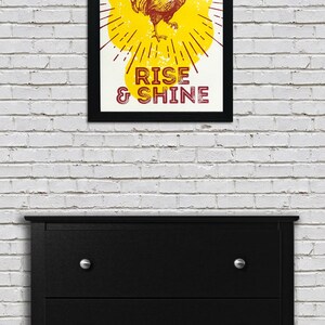 May include: A framed print with a yellow background and a rooster illustration. The rooster is red and has a yellow comb. The text "RISE & SHINE" is printed in red below the rooster.
