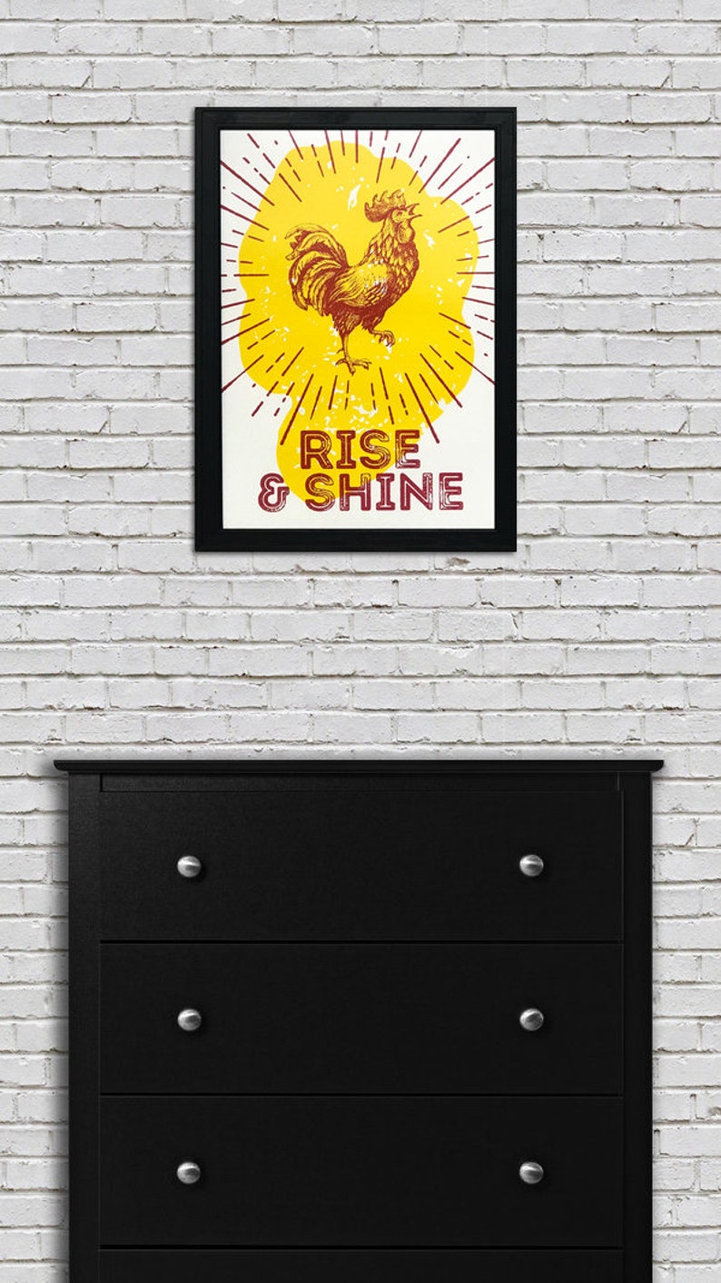 May include: A framed print with a yellow background and a rooster illustration. The rooster is red and has a yellow comb. The text "RISE & SHINE" is printed in red below the rooster.