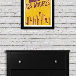 May include: A framed poster with a yellow background and the words "LOS ANGELES" in a distressed font. A basketball graphic is above the text. Below the text is a purple skyline silhouette. The frame is black.