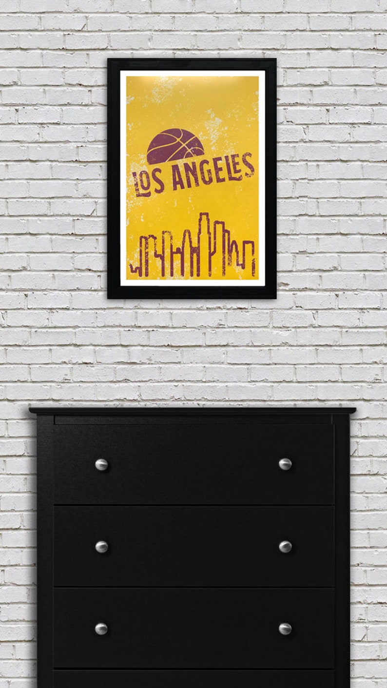 May include: A framed poster with a yellow background and the words "LOS ANGELES" in a distressed font. A basketball graphic is above the text. Below the text is a purple skyline silhouette. The frame is black.