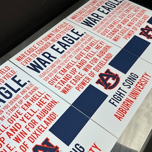 K&ouml;nnte beinhalten: Ein wei&szlig;es Schild mit blauem und rotem Text und einem blau-roten Logo. Der Text lautet "War Eagle, Fly Down the Field, Ever to Conquer, Never to Yield." Das Logo ist ein "A" mit einem roten "A" auf einem blauen "A". Der Text lautet auch "Give 'em Hell, Give 'em Hell, Stand Up and Yell, Hey! War Eagle, Win for Auburn, Power of Dixieland!" und "Fight Song, Auburn University".