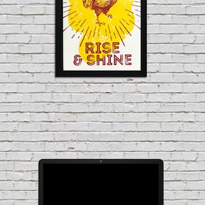May include: A framed print with a yellow background and a red rooster illustration. The rooster is crowing and has a sunburst behind it. The text "RISE & SHINE" is below the rooster.
