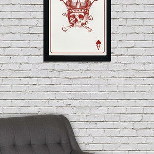 May include: Framed art print featuring a red skull wearing a crown, with crossbones and the Ace of Hearts and Spades symbols. The artwork is set against a white background and framed in black. The print is displayed on a white brick wall.