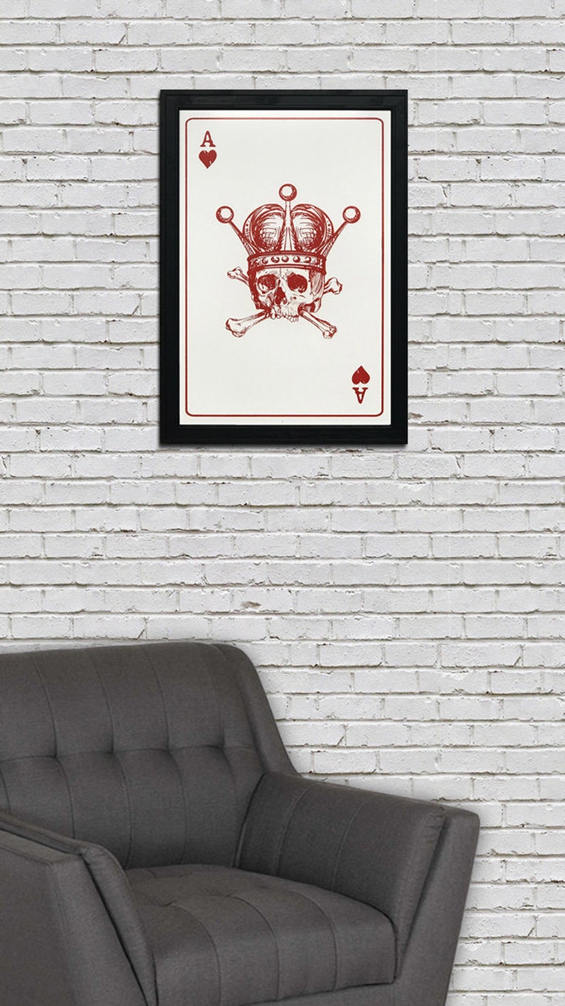 May include: Framed art print featuring a red skull wearing a crown, with crossbones and the Ace of Hearts and Spades symbols. The artwork is set against a white background and framed in black. The print is displayed on a white brick wall.