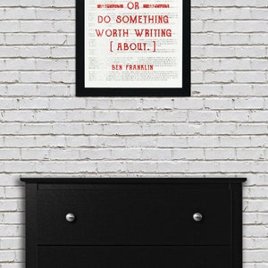May include: A black framed print with a white background and red text that reads "EITHER WRITE SOMETHING WORTH READING OR DO SOMETHING WORTH WRITING [ABOUT.] BEN FRANKLIN". The print is hanging on a white brick wall above a black dresser with four drawers.