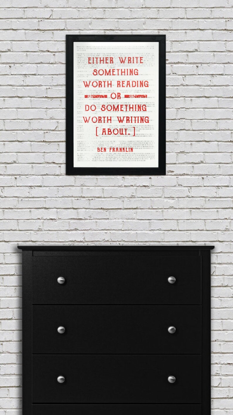 May include: A black framed print with a white background and red text that reads "EITHER WRITE SOMETHING WORTH READING OR DO SOMETHING WORTH WRITING [ABOUT.] BEN FRANKLIN". The print is hanging on a white brick wall above a black dresser with four drawers.