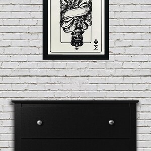 May include: A black and white illustration of the King of Clubs playing card. The King is depicted as a skeleton wearing a crown and wrapped in a banner. The card is framed in black.