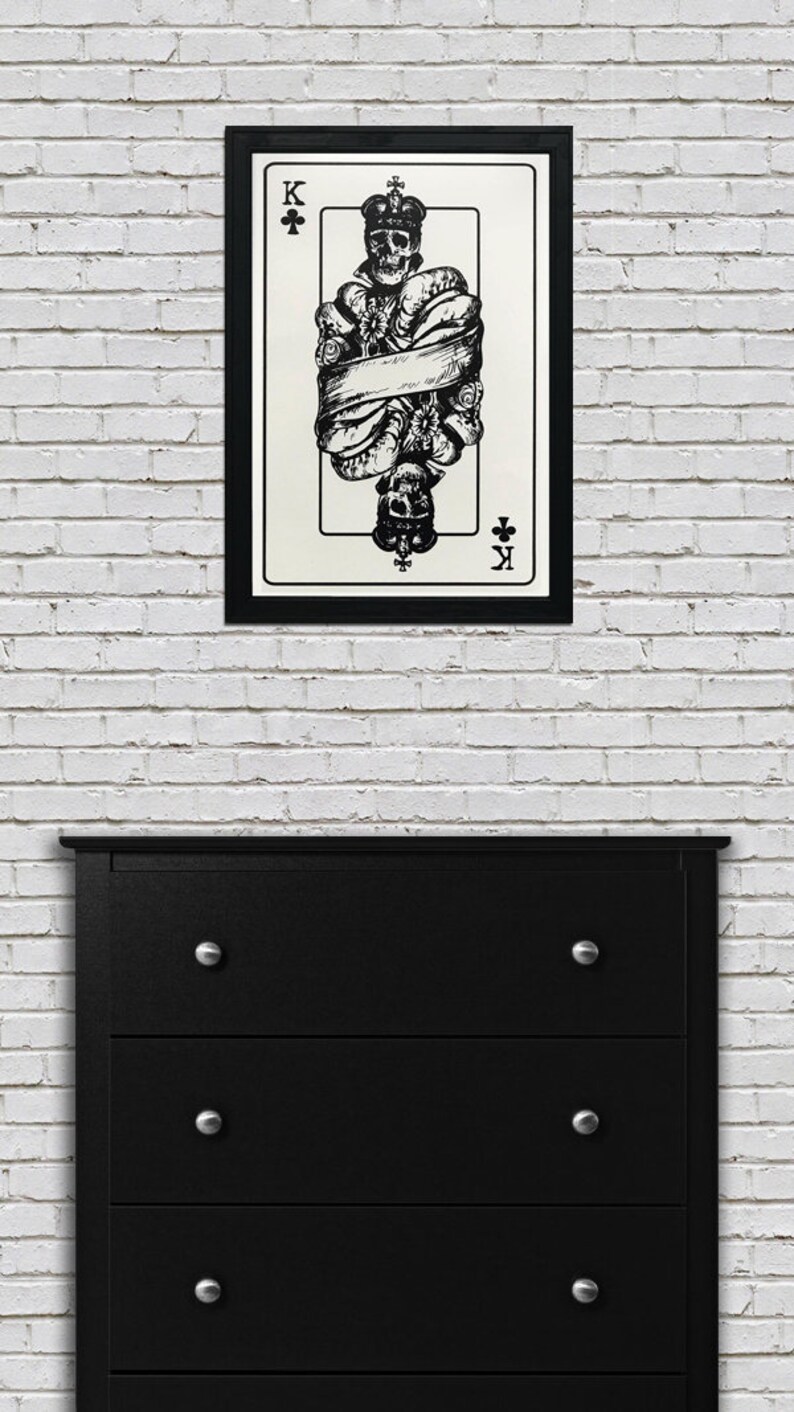 May include: A black and white illustration of the King of Clubs playing card. The King is depicted as a skeleton wearing a crown and wrapped in a banner. The card is framed in black.