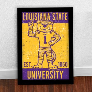 May include: Framed print with the Louisiana State University tiger mascot in a number 1 jersey. The print has a distressed, vintage style, with the text "LOUISIANA STATE UNIVERSITY" and "EST. 1860". The colours are purple and gold.