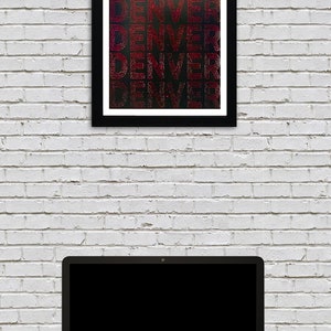 May include: A framed print with the word "DENVER" repeated five times in a red, white, and blue color scheme. The print is on a black background and has a white brick wall behind it. A laptop computer is in the foreground.