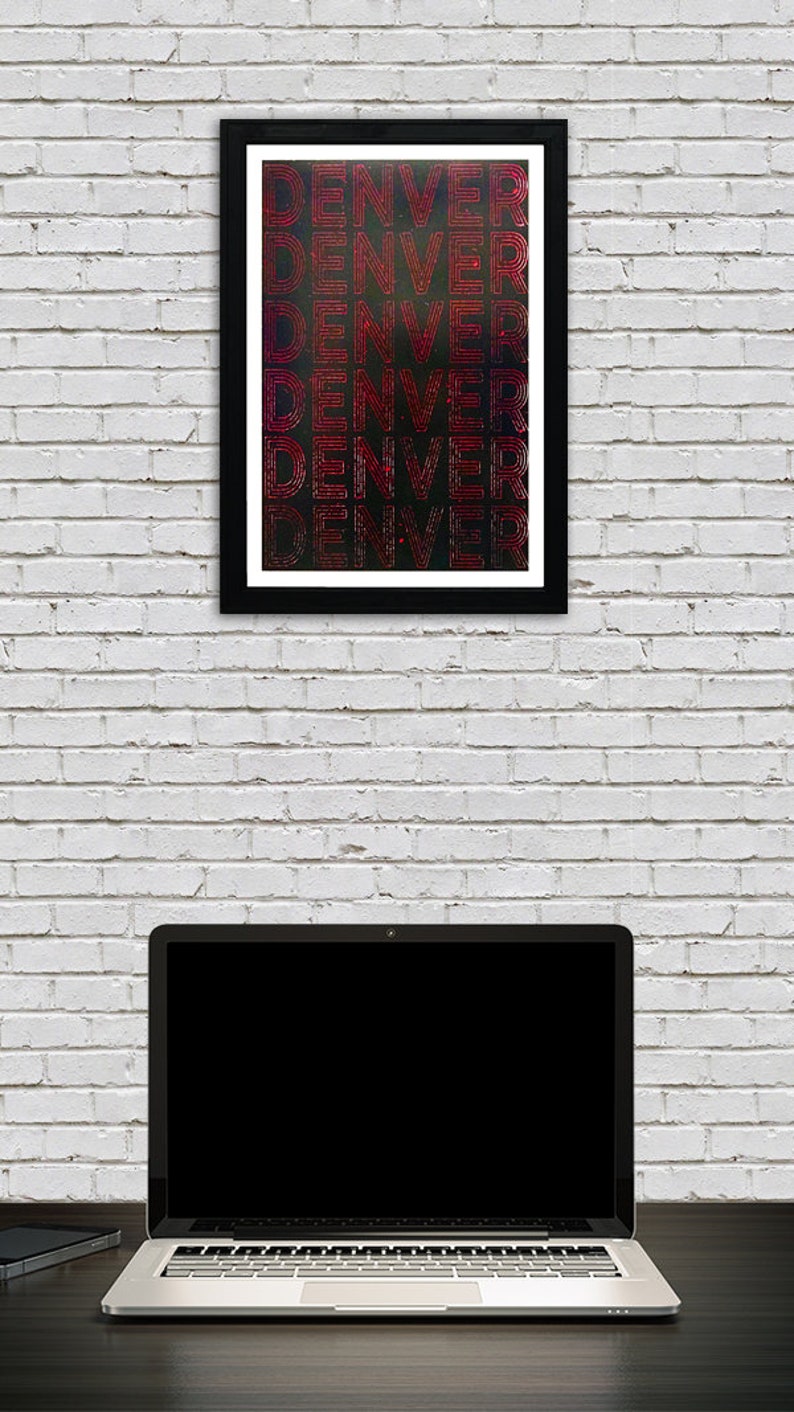 May include: A framed print with the word "DENVER" repeated five times in a red, white, and blue color scheme. The print is on a black background and has a white brick wall behind it. A laptop computer is in the foreground.