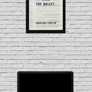 May include: A framed black and white print with the quote "The ballot is stronger than the bullet. - Abraham Lincoln" on a white brick wall with a laptop computer on a dark wood surface.