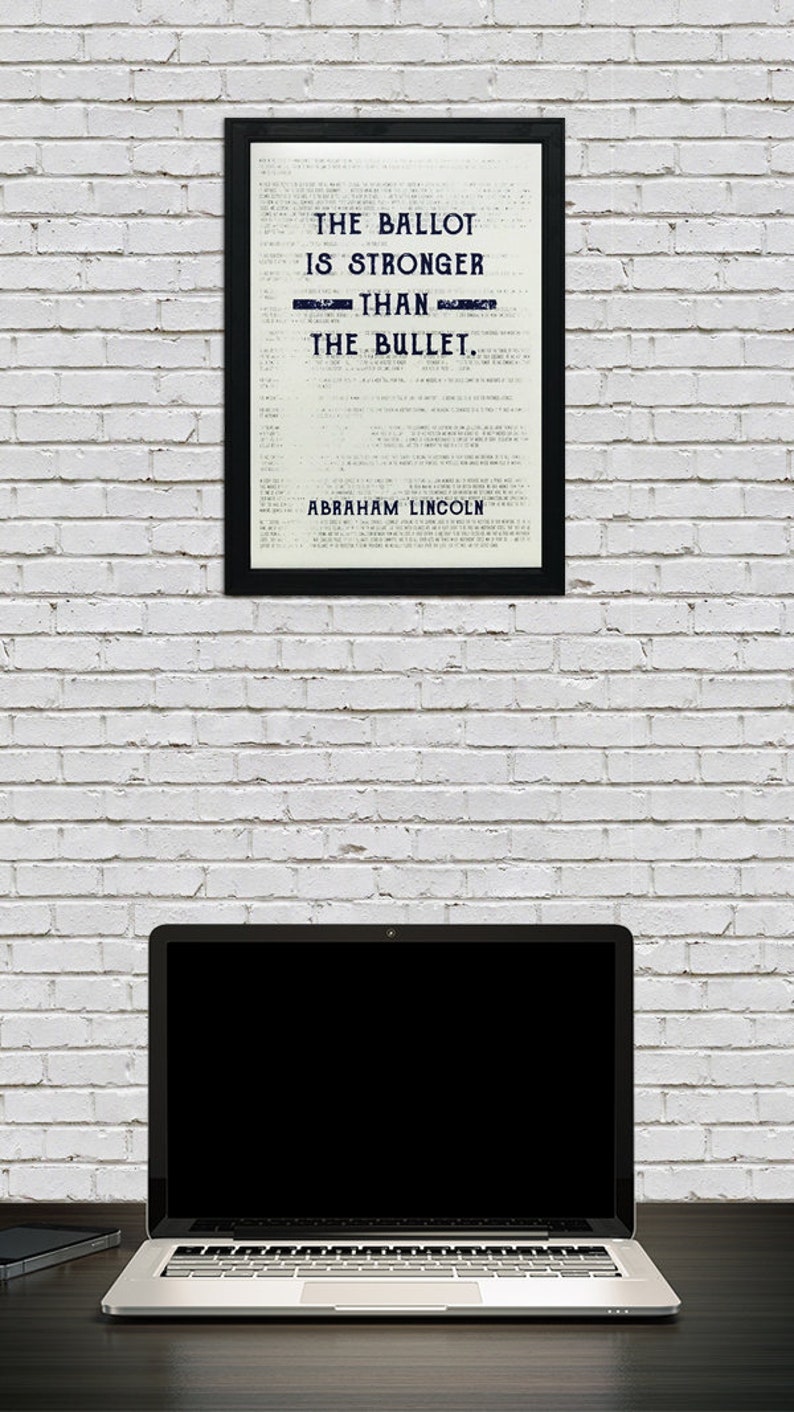 May include: A framed black and white print with the quote "The ballot is stronger than the bullet. - Abraham Lincoln" on a white brick wall with a laptop computer on a dark wood surface.
