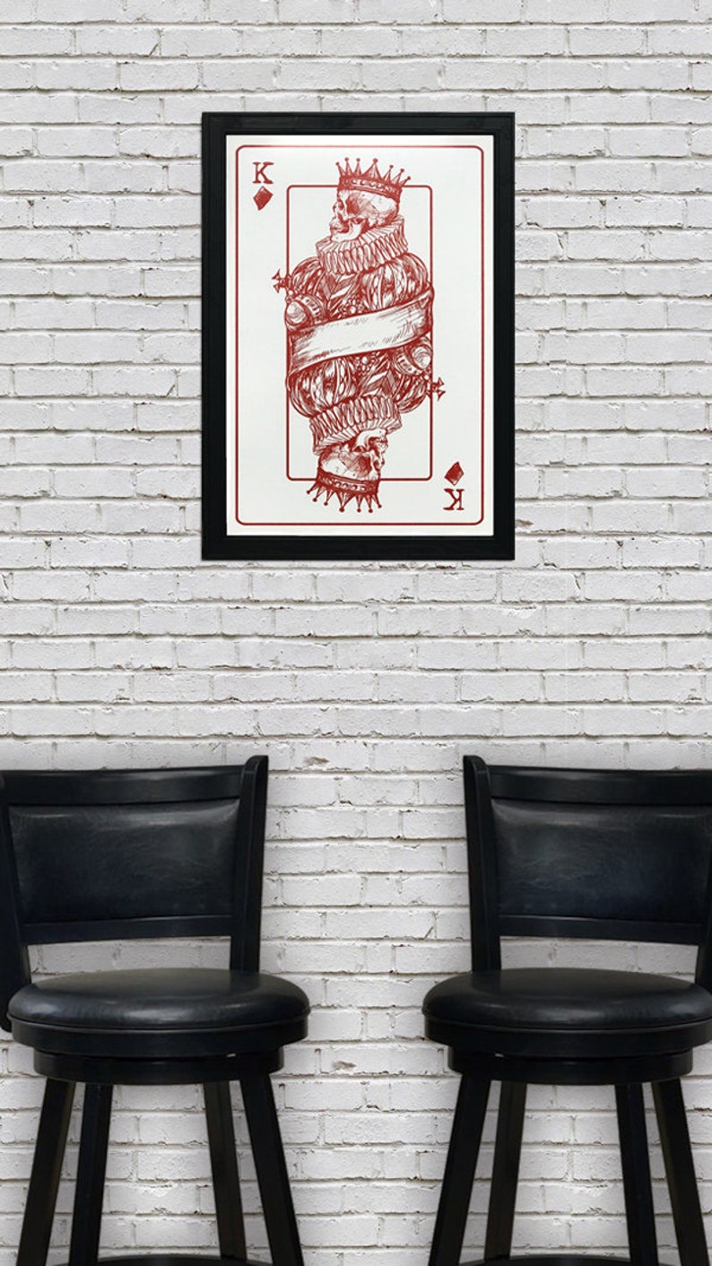 May include: A framed print of a red and white playing card featuring the King of Diamonds. The card depicts a skull wearing a crown and a ruff collar.