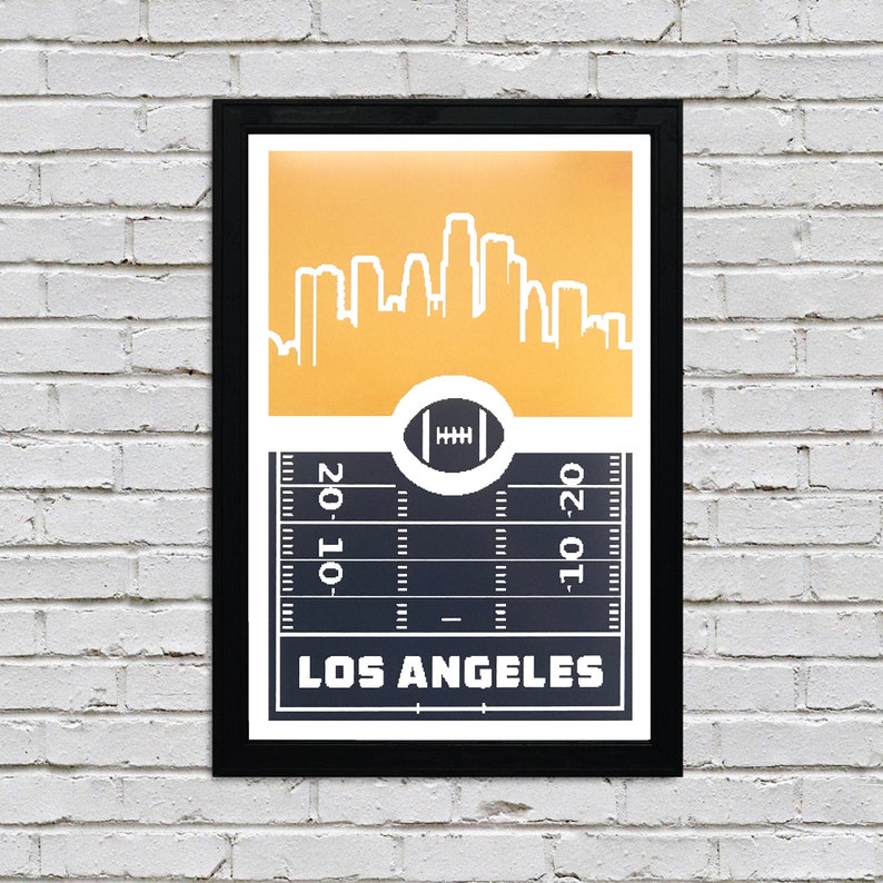 May include: A black framed print with a gold background featuring a white silhouette of the Los Angeles skyline. The bottom of the print features a black and white football field with the words "Los Angeles" printed in white.