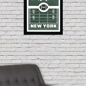 Puede incluir: Una impresi&oacute;n enmarcada con un fondo verde que presenta una silueta blanca del horizonte de la ciudad de Nueva York. El horizonte se coloca sobre un campo de f&uacute;tbol con l&iacute;neas de yarda y el texto "NEW YORK" debajo.
