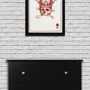 May include: Framed art print featuring a red illustration of a skull wearing a crown and crossed bones, set against a white background. The artwork is the Ace of Spades card. The frame is black, and the print hangs on a white brick wall.