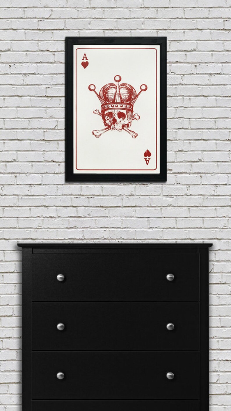 May include: Framed art print featuring a red illustration of a skull wearing a crown and crossed bones, set against a white background. The artwork is the Ace of Spades card. The frame is black, and the print hangs on a white brick wall.