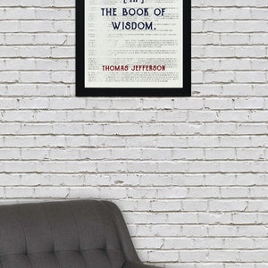May include: A framed print with the quote "HONESTY IS THE FIRST CHAPTER [IN] THE BOOK OF WISDOM" by Thomas Jefferson. The text is in black and blue on a white background, framed in black. A gray armchair is in the foreground.