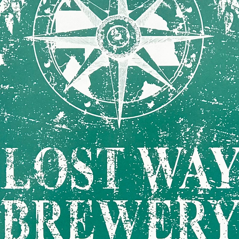 May include: A distressed teal green sign with white text that reads "Lost Way Brewery". A compass with a white background and black lines is in the upper left corner of the sign.