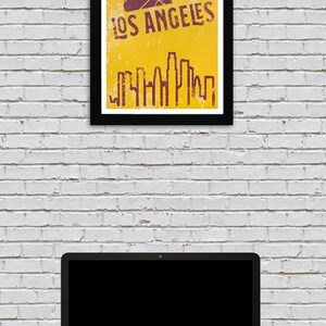 May include: A framed poster with a black border featuring a basketball and the words "LOS ANGELES" in a distressed font. Below the text is a skyline silhouette. The poster is against a white brick wall, with a laptop and phone on a desk.
