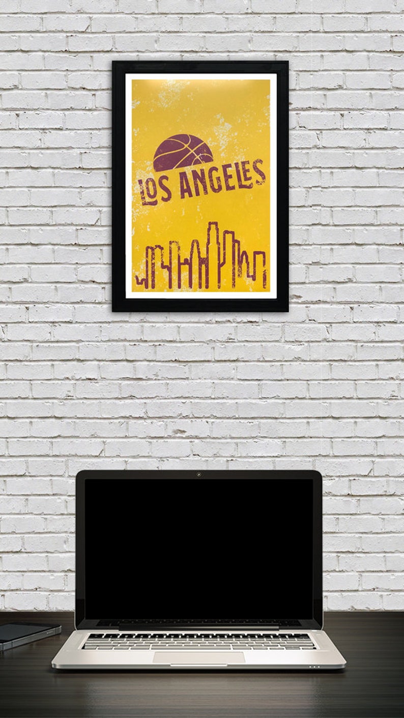 May include: A framed poster with a black border featuring a basketball and the words "LOS ANGELES" in a distressed font. Below the text is a skyline silhouette. The poster is against a white brick wall, with a laptop and phone on a desk.