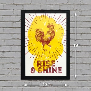 May include: A black framed poster with a yellow and red rooster illustration. The rooster is crowing and has a sunburst behind it. The text "Rise & Shine" is printed in red below the rooster.