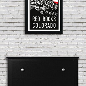 May include: A black and white illustration of Red Rocks Amphitheatre in Morrison, Colorado. The illustration is set against a red and white sunburst background. The text "RED ROCKS COLORADO" is printed below the illustration.