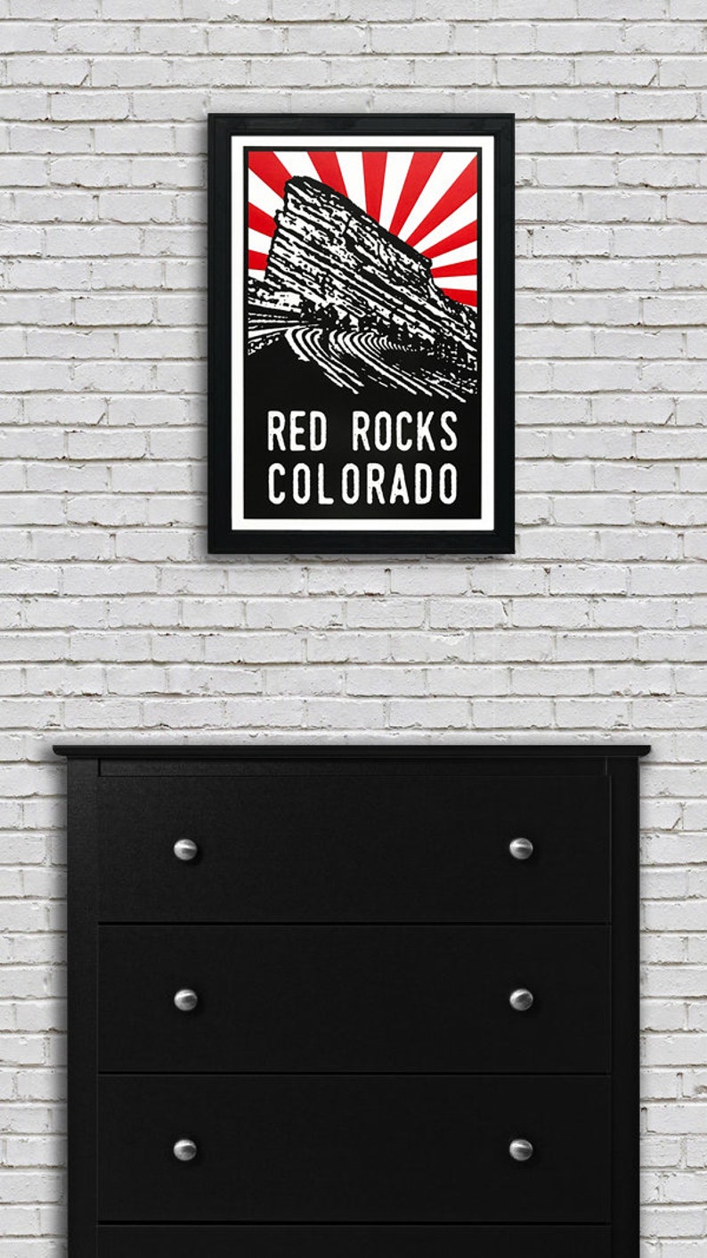 May include: A black and white illustration of Red Rocks Amphitheatre in Morrison, Colorado. The illustration is set against a red and white sunburst background. The text "RED ROCKS COLORADO" is printed below the illustration.