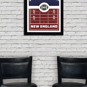 May include: A framed print with a navy blue background featuring a white silhouette of a city skyline. The bottom of the print has a red and white football field design with the text "NEW ENGLAND" and yard lines marked "20", "10", and "10-20".