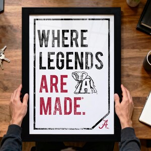 A person is holding up a framed poster with the text "WHERE LEGENDS ARE MADE" and an illustration of an elephant.