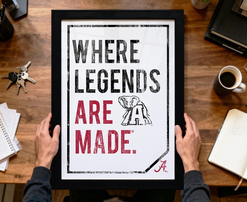 A person is holding up a framed poster with the text "WHERE LEGENDS ARE MADE" and an illustration of an elephant.