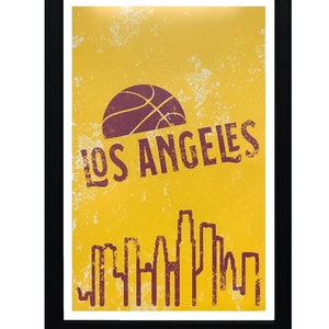 May include: A framed poster with a yellow background and a distressed texture. The poster features a basketball graphic, the text "LOS ANGELES", and a skyline silhouette in a dark red color. The frame is black.